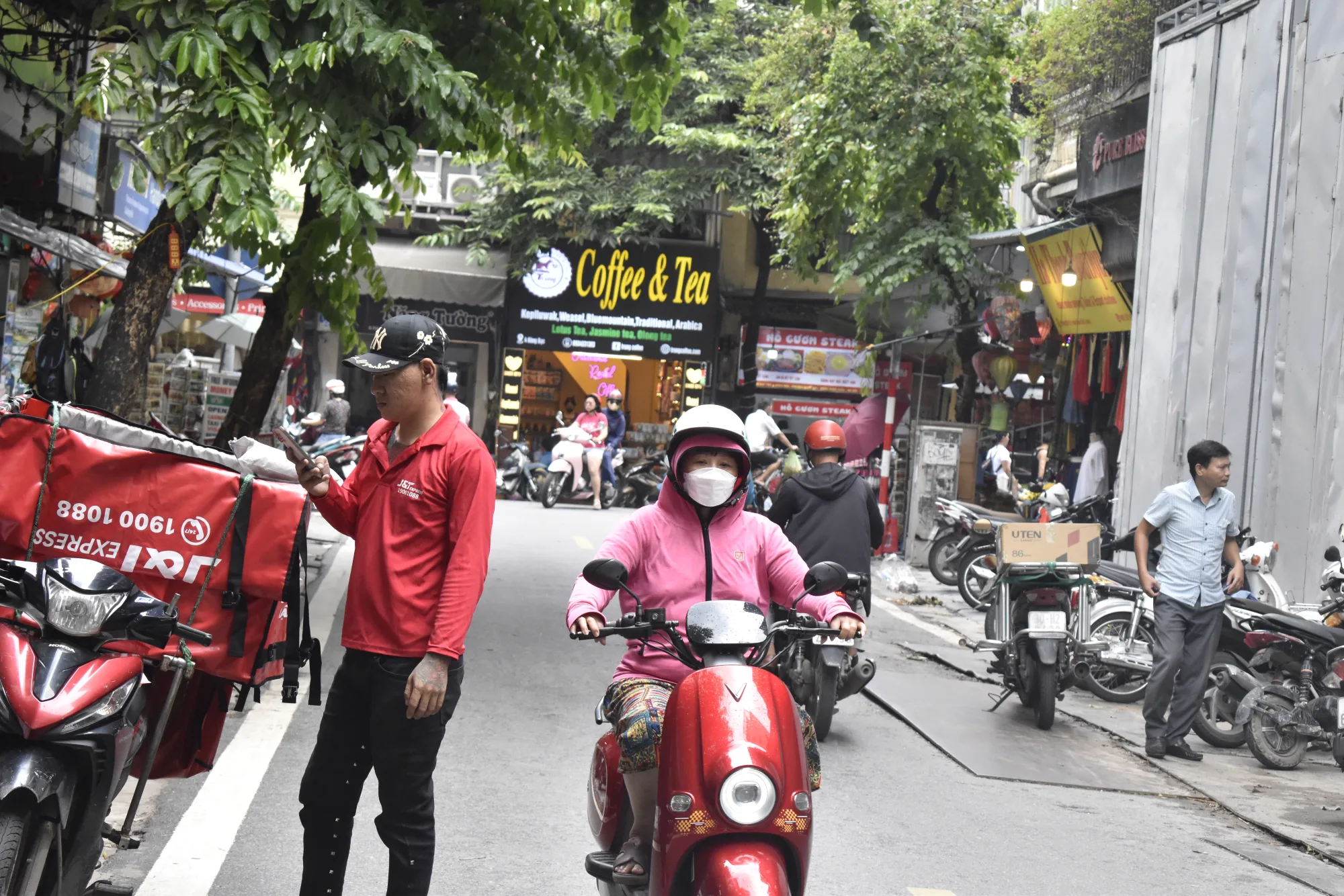 A street in Hanoi's Old Quarter with scooters, delivery riders, and cafe signboards